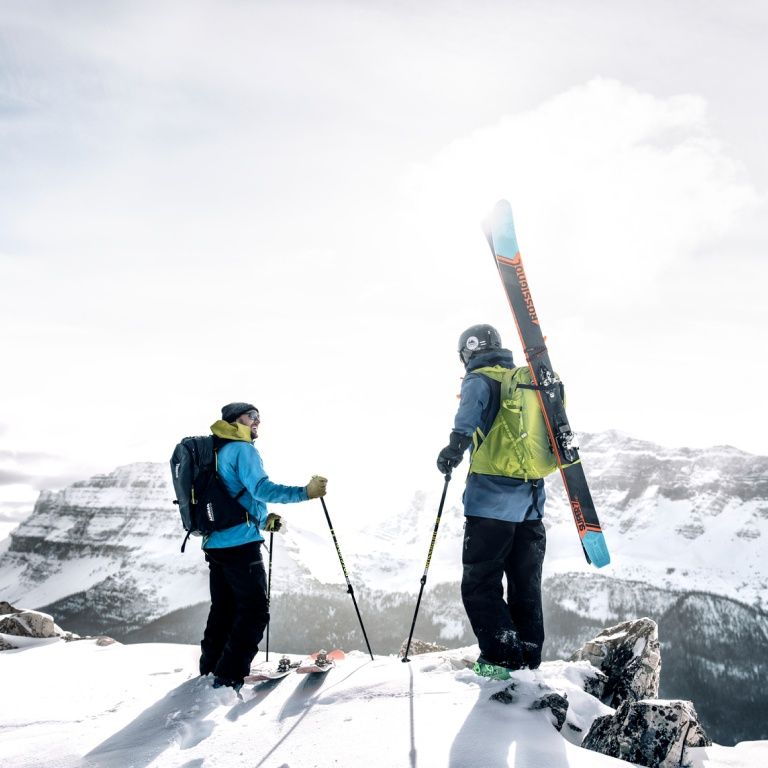Two people stand on a mountain with skis and ski backpacks.