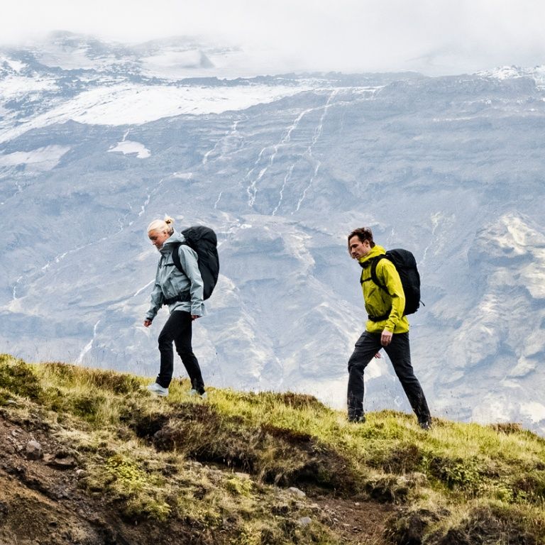 Two people walk on a wooden trail in a field carrying Thule hiking backpacks.