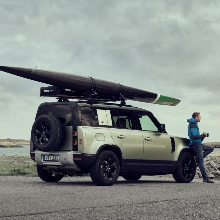 A man holding a mug leans against the front of his SUV which is equipped with a closed Thule rooftop tent and Thule kayak rack holding a kayak.
