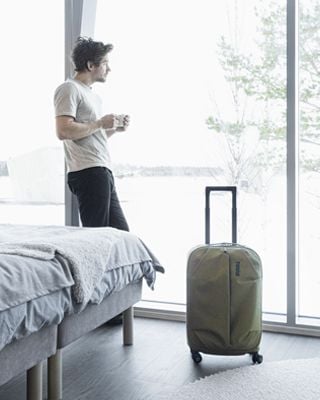 A man in a hotel room looks out the window with a coffee and a suitcase beside him.