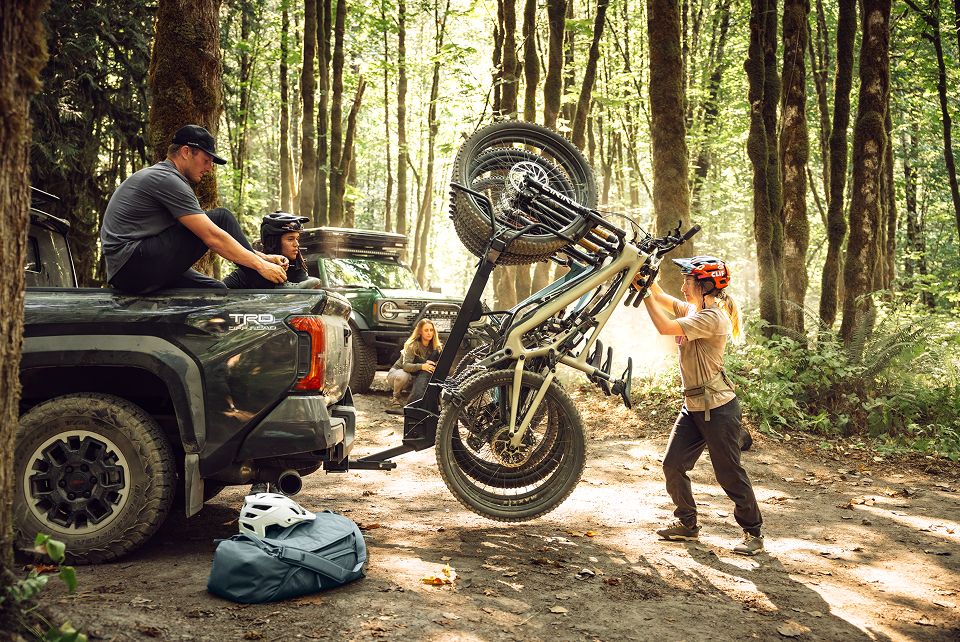 A group of mountain bikers gather around a pickup truck in a dense forest, with one rider lifting a bike onto a vertical Thule ReVert hitch rack, while others relax nearby.