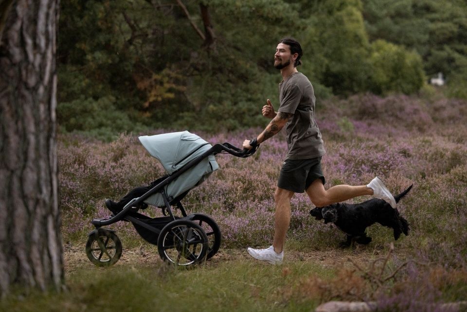 A man pushes a Thule Urban Glide 3 stroller with a child inside while running in nature with his dog.