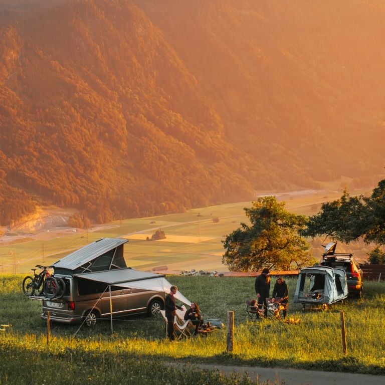 People sit in chairs next to two vehicles equipped with a Thule rooftop tents and Thule Outset hitch tent in nature.