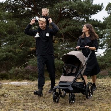 A man with a toddler sitting on his shoulders walks in nature next to a woman pushing an empty Thule Urban Glide stroller.