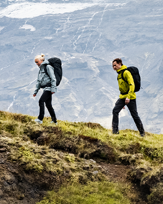 Two people walk on a wooden trail in a field carrying Thule hiking backpacks.