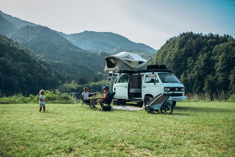 A family is sitting outside a vintage Volkswagen van with a Thule Approach rooftop tent in the Austrian alps.