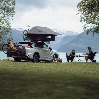 Two people sit in folding chairs at a folding table next to a car equipped with a Thule rooftop tent and Thule towbar bike rack.