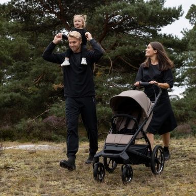 A man with a toddler sitting on his shoulders walks in nature next to a woman pushing an empty Thule Urban Glide stroller.