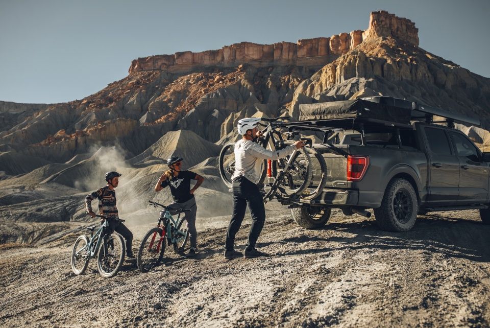 A man unloads a bike from the Thule Verse hitch mounted bike rack while it's attached to the back of a truck and two people sitting on bikes look on.